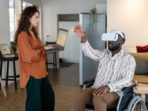 A woman on a computer interacts with a man in a wheelchair waring XR (VR) goggles