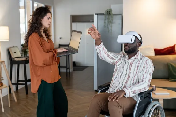 A woman on a computer interacts with a man in a wheelchair waring XR (VR) goggles